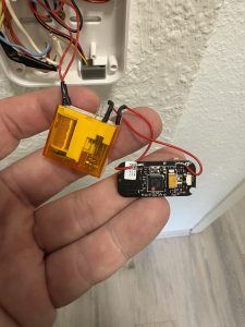 Nahaufnahme einer geöffneten Türklingel- oder Gegensprechanlage. Eine Hand hält eine kleine elektronische Platine mit angelötetem gelbem Relais und roten Drähten.
