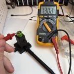 Ein Multimeter welches 1,302V anzeigt. Im Vorgerund eine Hand die eine Messspitze auf den Pluspol eines AA Akkus hält welcher in einer Halterung sitzt. Unten in der Halterung steckt die zweite Messspitze