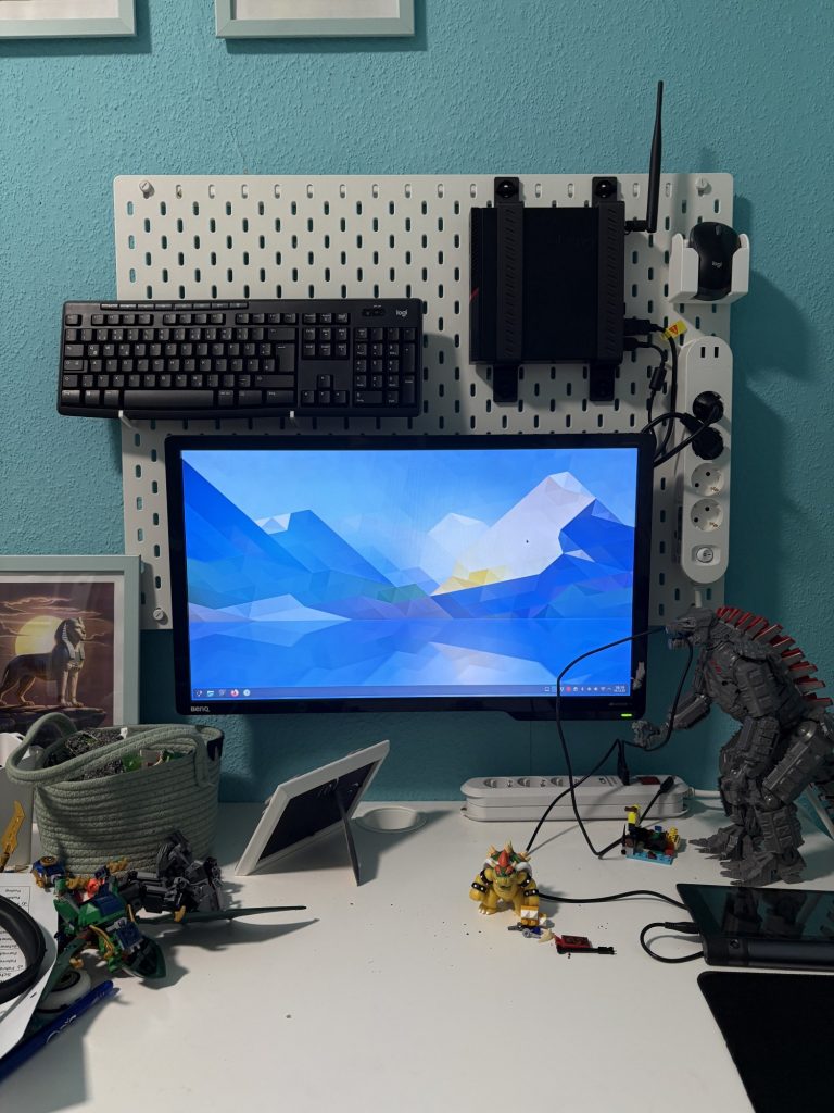 Das Bild zeigt einen Arbeitsbereich mit einem weißen Schreibtisch vor einer hellblauen Wand. Monitor und Ausstattung: Auf dem Schreibtisch steht ein großer BenQ-Monitor, auf dessen Hintergrundbild eine abstrakte, farbenfrohe Berglandschaft zu sehen ist. Über dem Monitor befindet sich eine weiße Stecktafel, an der eine schwarze Tastatur (Logitech) und ein schwarzer Mini-PC befestigt sind. Rechts neben dem Monitor ist eine weiße Steckdosenleiste mit mehreren Steckern angebracht. Dekorationen und Spielzeug: Der Schreibtisch ist mit mehreren Lego- und Actionfiguren dekoriert, darunter ein gelbes Fantasiewesen und ein großer Roboter. Auf der linken Seite befindet sich ein Korb mit Kabeln und ein kleines Bild mit einer ägyptischen Sphinx. Zubehör: Vor dem Monitor steht ein weißer Bilderrahmen. Rechts davon befindet sich ein schwarzes Computer-Mauspad.