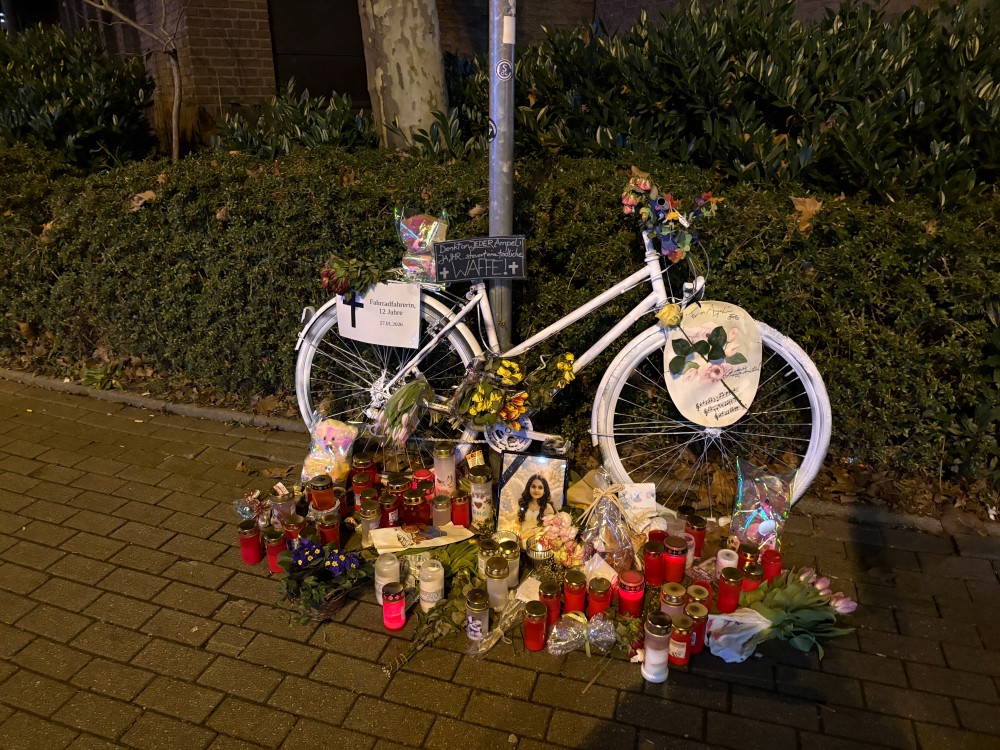 Nachtfoto eines an einen pfosten gelehnten Ghostbikes vor einer Hecke. Davor Blumen, Grabkerzen, Spielzeuge und das Bild eines Mädchens in festlicher Kleidung mit Engelflügeln scheinbar vor den Himmelstoren. Es gibt mehrere Schilder am Fahrrad.Auf einem ist ein Kreuz zu sehen, daneben die Aufschrift: Fahrradfahrerin, 12 Jahre 27.02.2026. Auf einem weiteren kann man lesen: Denkt an jeder Ampel: ja Ihr steuert eine tötliche Waffe!