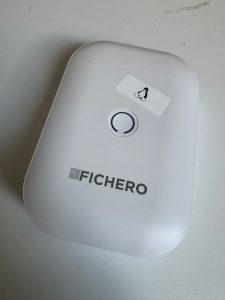 Ein weißes rechteckiges Gerät mit abgerundeten Kanten liegt auf einer weißen Tischplatte. In der Mitte ein Runder Knopf darunter die Aufschrift Fichero. Über dem Knopf eine Klebeetikett mit Tux dem Linux Kernel Symbol.