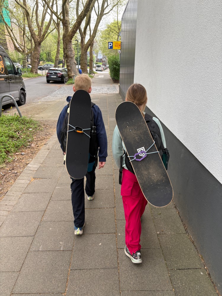 Zwei Kinder laufen auf einem Gehweg. Beide haben Skateboards auf dem Rücken. Ein Kind trägt eine blaue Hose das andere eine rote.