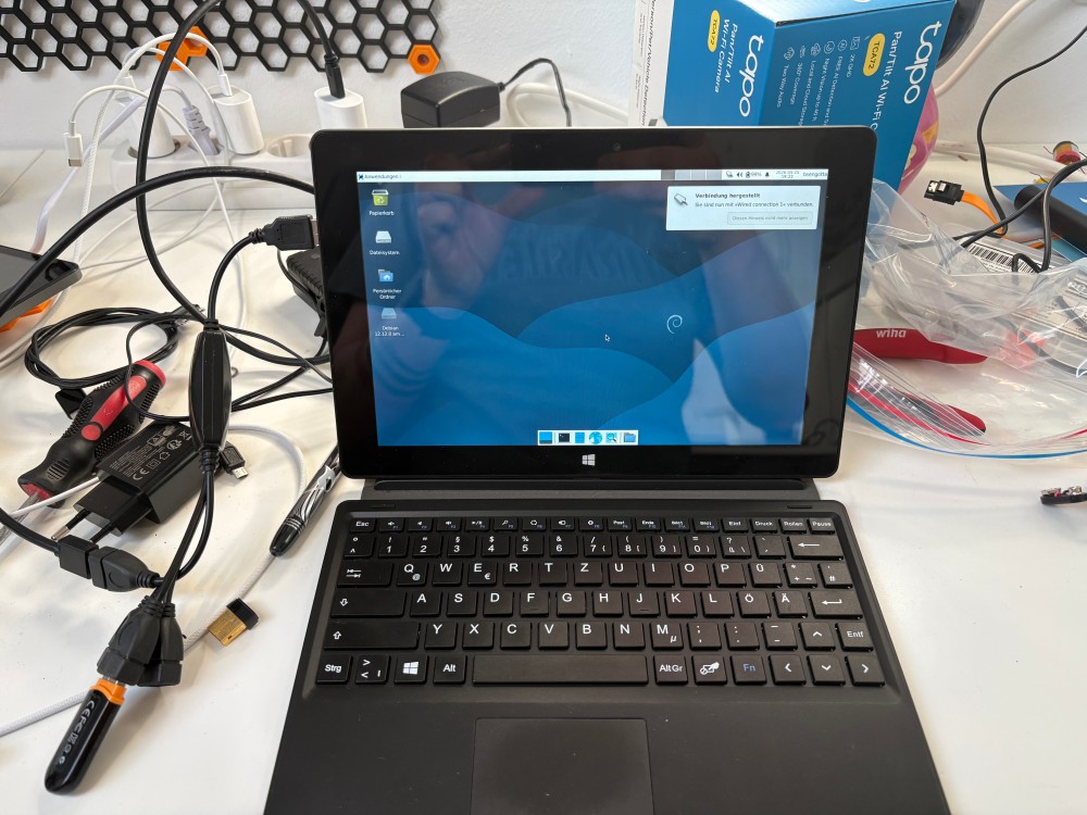 Ein Laptop auf dem Debian 13 XFCE läuft, Desktop ist sichtbar, rundherum liegt Werkzeug im Hintergrund ist eine Steckdosenleiste zu erkennen