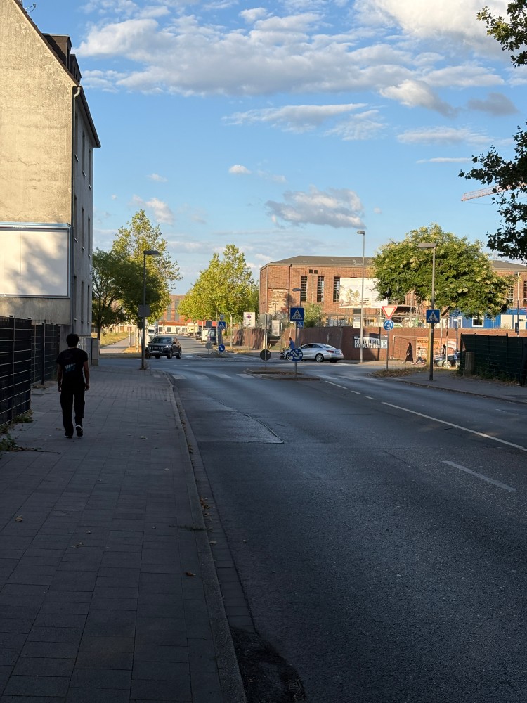 Eine Strasse fotografiert vom Gehweg aus. Man sieht zwei Autos im Kreisverkehr sowie einige Passanten auf den Gehwegen. Im Hintergrund kann man zwei Gebäude erkennen die zum Schulgelände gehören.